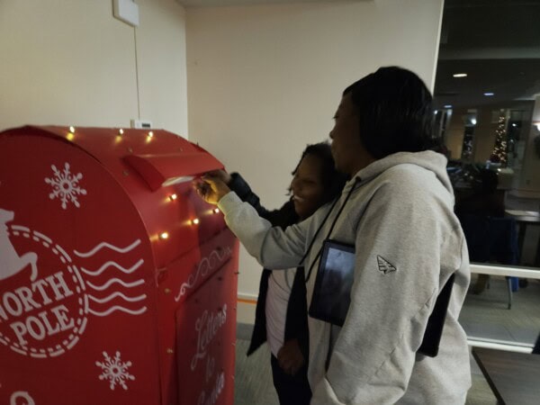 Two women are smiling as they place a letter into a large red mailbox decorated with holiday lights and labeled North Pole inside a building.