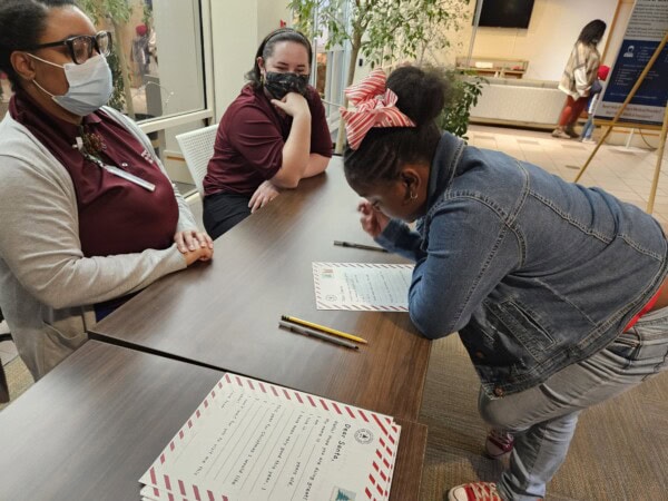 A young girl leans over a table to fill out a form while two adults, one wearing a mask, sit across from her. Pencils and more forms are placed on the table in a brightly lit room.