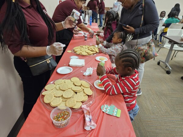 Children decorate cookies at a long table covered with a red tablecloth. Various toppings and plain cookies are visible. Adults assist and supervise, and the room has a warm, lively atmosphere.