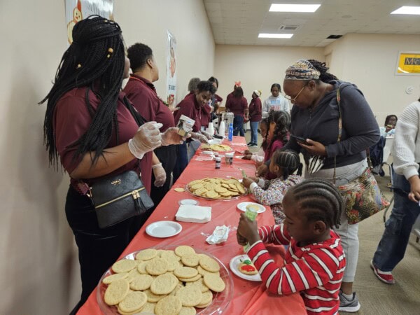 Children and adults gather around a long table covered with cookies and decorating supplies. Volunteers in matching shirts help kids decorate cookies at a community event in a brightly lit room.