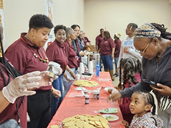 A group of people serves cookies and drinks at a long table covered with a red cloth, while children and adults enjoy snacks. The room appears lively and festive, with people interacting and smiling.