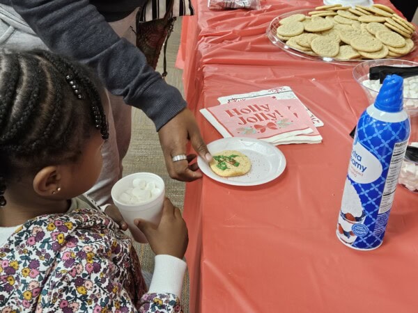 A child holds a cup of marshmallows while an adult decorates a cookie on a plate at a red table with cookies, napkins that say Holly Jolly!, and a can of whipped cream.