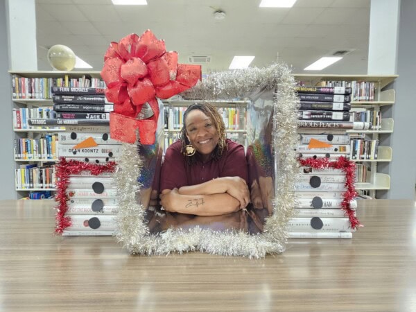 A smiling woman poses behind a festive, tinsel-framed display with a large red bow, surrounded by stacked books, in a library with bookshelves in the background.