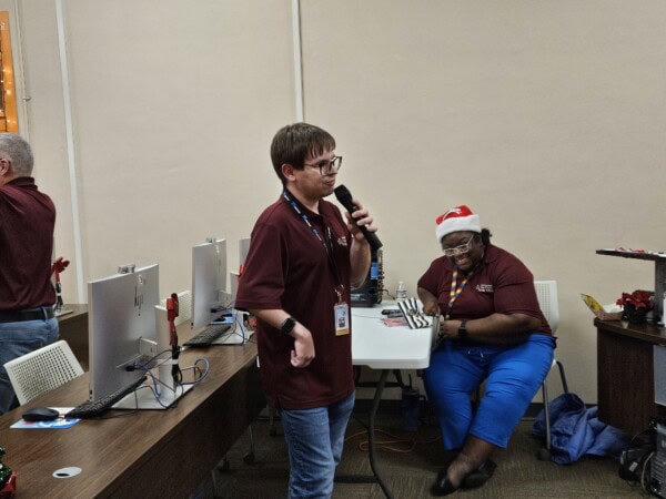 A person stands speaking into a microphone in an office with computers, while another person wearing a Santa hat sits and smiles nearby. Both are wearing maroon shirts and name badges.