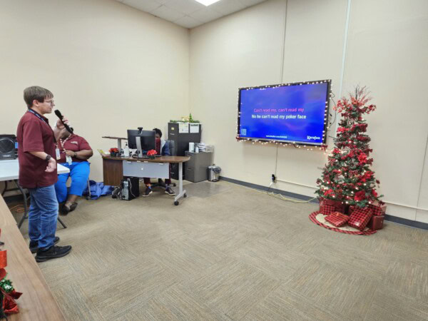A person sings karaoke while reading lyrics on a large screen in an office decorated with a Christmas tree and wrapped presents. Two other people sit by a desk with computers in the background.