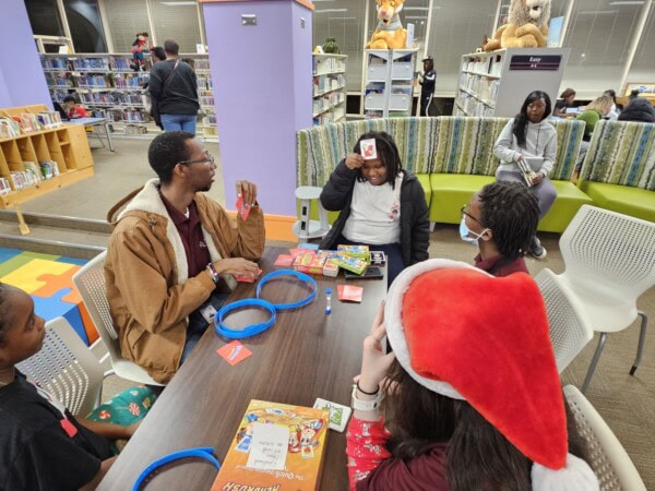 Four people sit around a table playing a guessing game with headbands in a library. One wears a Santa hat, another holds a card to their forehead. Bookshelves and large stuffed animals are in the background.