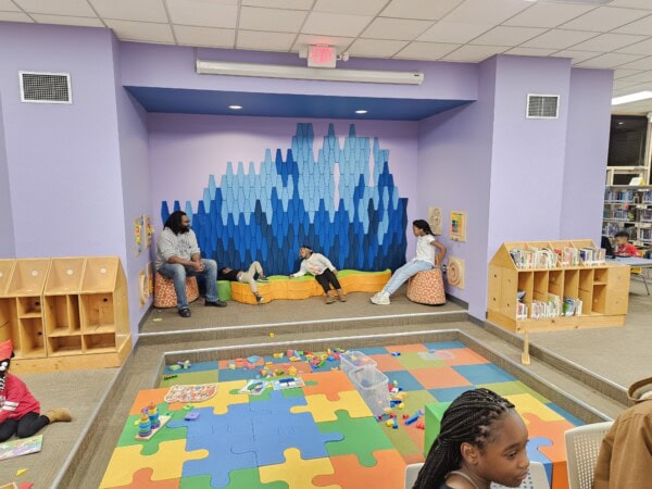 Children and adults sit and play in a colorful library reading nook with a blue geometric wall, foam floor tiles, toys, and bookshelves. The atmosphere looks lively and welcoming.