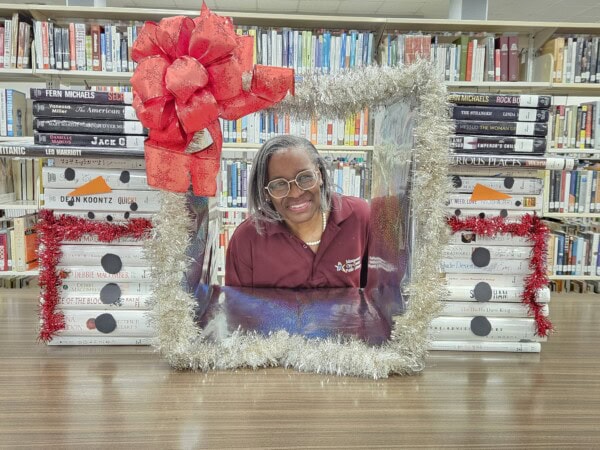 A smiling woman sits at a library table framed by silver tinsel with a large red bow. Behind her are stacks of books decorated with red tinsel and black circles, resembling festive holiday gifts. Shelves of books fill the background.
