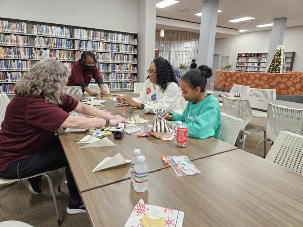 Four people sit around a table in a library doing arts and crafts with paper and glue. Library shelves filled with books are in the background, and a decorated Christmas tree is visible on the right.