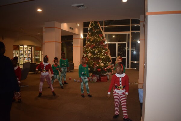 Seven children dressed in festive elf and Santa outfits stand and pose near a decorated Christmas tree with presents underneath, inside a brightly lit room with holiday decorations.
