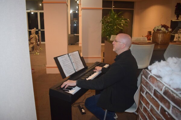 A bald man in glasses and a black jacket plays an electric keyboard with sheet music in a warmly lit indoor setting, seated near a brick wall and plants.