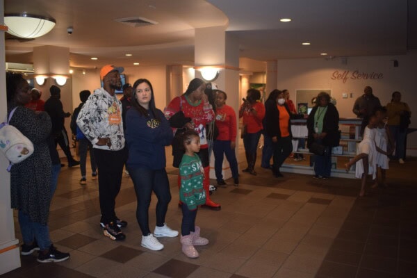 A diverse group of adults and children stand in a lobby area, some watching something off-camera. The atmosphere is casual, with people in sweaters and jackets. Two young girls in festive outfits stand near the center.
