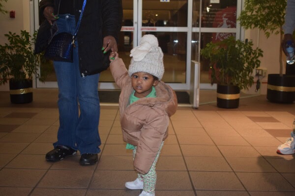 A small child in a beige coat and knit hat holds an adult’s hand indoors, looking at the camera. The adult wears jeans and a dark jacket; only their lower body is visible. Plants and glass doors are in the background.