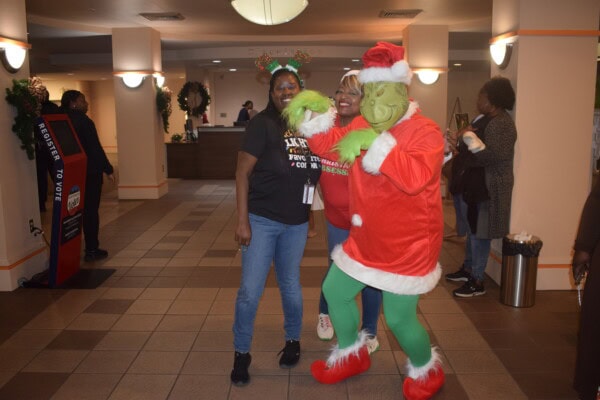 Three people pose indoors; one wears reindeer antlers, another wears a red top, and the third is dressed as the Grinch in a Santa suit. Holiday decorations are visible in the background.