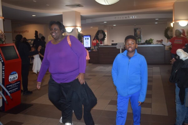 A woman in a purple top and a boy in a blue tracksuit walk through a lobby with tiled floors. Other people are in the background near a circulation desk decorated with wreaths.