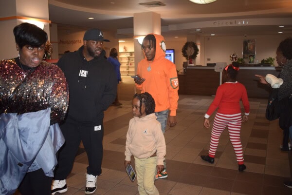 A group of people, including children and adults, walk through a library lobby. Some are looking around, and one man checks his phone. A girl in red and white stripes stands near the circulation desk decorated with a wreath.