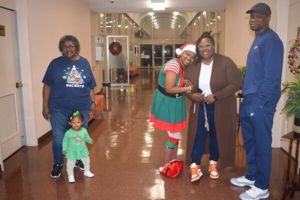 Five people pose in a hallway with holiday decorations. A woman in a Christmas sweater stands on the left with a toddler in green, while an adult dressed as an elf hugs another woman. A man in a blue tracksuit stands on the right.