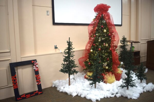 A decorated Christmas tree with red ribbon and ornaments stands among small artificial pine trees and fake snow. A black frame labeled MCPL FAMILY GAME NIGHT leans on the left side.