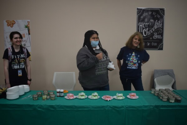 Three women stand behind a green table with jars and holiday treats. One wears a mask on her chin, another has a festive sweater, and a sign behind them reads Hot Chocolate Bar. They appear to be at a holiday event.