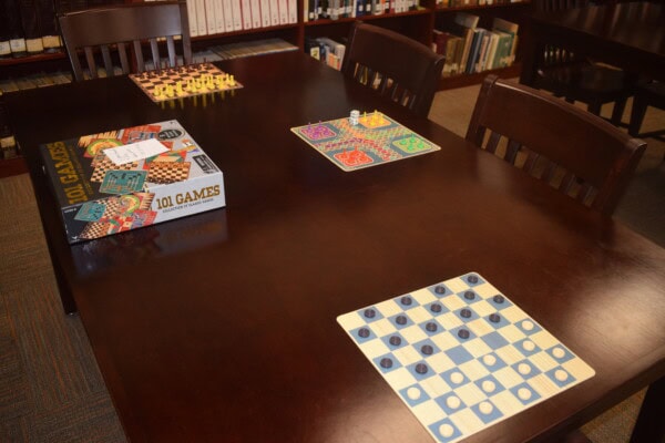 A wooden table in a library displays various board games, including checkers, chess, and a colorful card game. A “101 Games” box sits on the table, and bookshelves are visible in the background.
