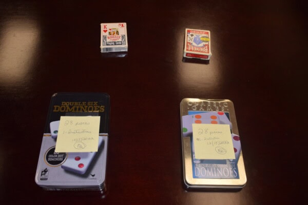 Four game sets—a small pack of playing cards, a full-size pack of playing cards, and two tins of dominoes—are arranged on a dark wooden table, each domino tin labeled with a handwritten sticky note.