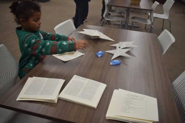 A child in a green sweater folds book pages into star shapes at a wooden table. There are open book pages, finished paper stars, and tape dispensers on the table. White chairs and another person are in the background.