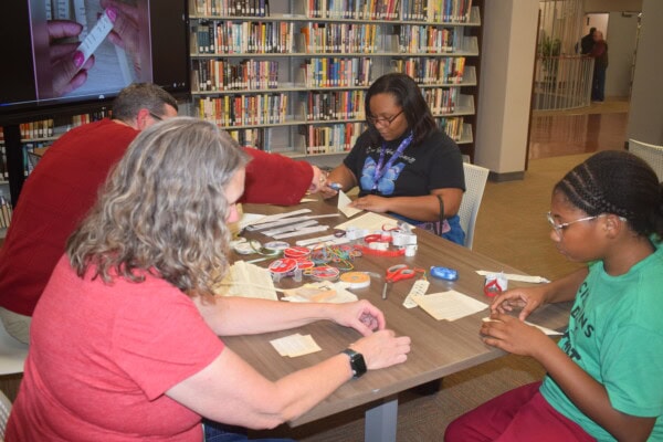 Four people sit around a library table, working on crafts with paper, ribbons, tape, and scissors. Bookshelves and another person are visible in the background. A screen displays an image of hands folding paper.