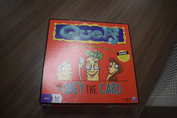 A red box of the party game Quelf sits on a wooden surface. The box features cartoon characters and the tagline Obey the Card. A yellow warning label and game details are also visible.