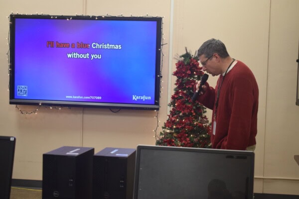 A man in a red sweater sings karaoke while looking at a screen displaying the lyrics Ill have a blue Christmas without you. A small decorated Christmas tree stands next to him in the room.