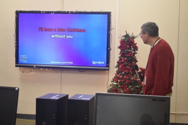 A man in a red sweater stands near a decorated Christmas tree, holding a microphone and looking at a screen displaying karaoke lyrics: Ill have a blue Christmas without you.
