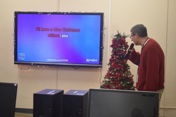 A man sings karaoke into a microphone next to a small decorated Christmas tree. The screen behind him displays lyrics from Blue Christmas with a festive background. Computer monitors are visible in the foreground.