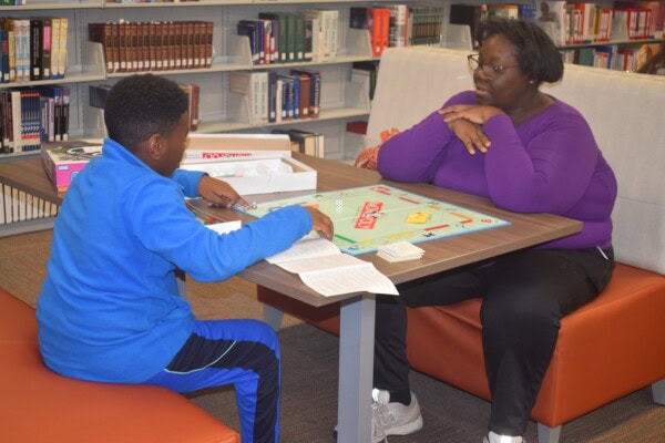 A boy and a woman sit across from each other playing Monopoly at a table in a library, surrounded by bookshelves. The boy is reading from the game instructions.