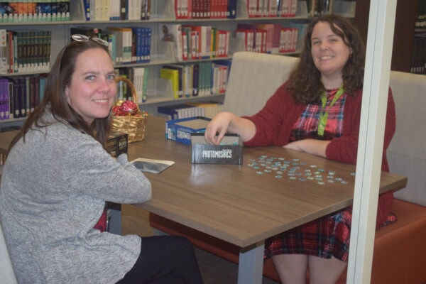Two women sit at a table in a library, smiling at the camera. They are working on a jigsaw puzzle together, with puzzle pieces spread out and a box labeled PROMOSIONES on the table. Bookshelves are in the background.
