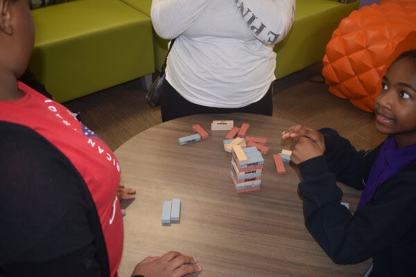 Three children play a game of Jenga at a round table, stacking and removing wooden blocks. One child is smiling, while another watches closely. A person in a white shirt stands nearby.