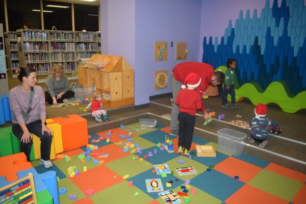 Children and adults play with colorful blocks and toys in a library play area. Some kids wear Santa hats, and library bookshelves are visible in the background. The room is bright and lively with playful decor.