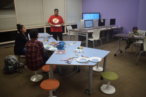 A woman stands and talks to two seated people at a table covered with crayons, coloring pages, and paper plates in a computer room, while a child sits alone at a desk in the background.