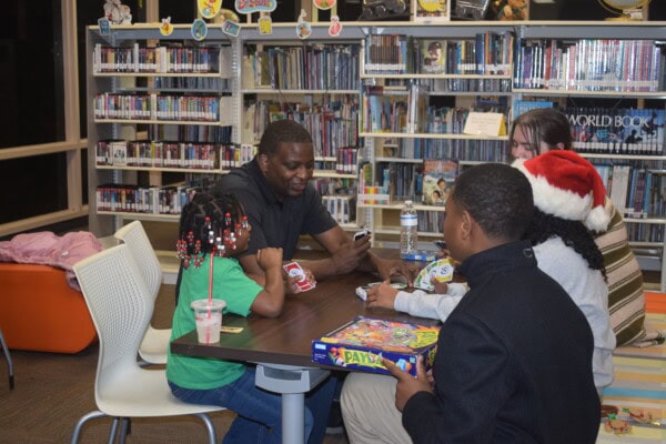 Four people sit around a table in a library playing board games. Shelves of books are in the background. One child wears a Santa hat, another has colorful beads in her hair, and there are drinks and game boxes on the table.