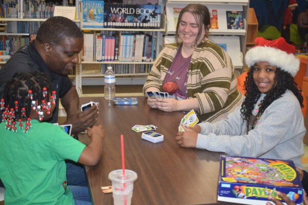 Four people sit around a table in a library playing an UNO card game. A girl in a Santa hat smiles at the camera. Board games and drinks are on the table; bookshelves are visible in the background.