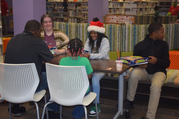 Four people sit around a table playing the card game UNO in a library. One person wears a Santa hat, another has red beads in their hair. A board game and a drink are on the table. Bookshelves are in the background.