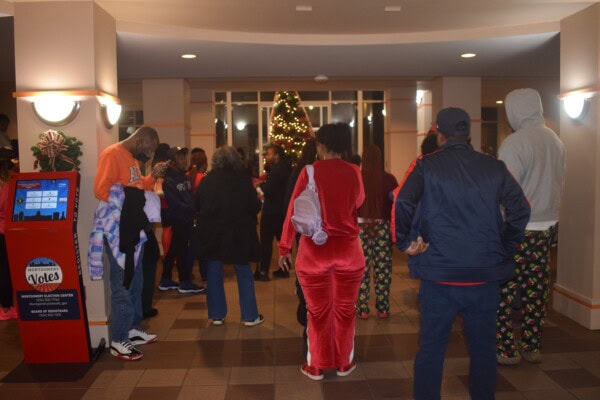 A group of people in festive pajamas gather in a decorated lobby with a lit Christmas tree in the background. A red kiosk stands on the left side of the image.