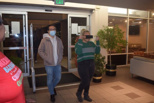 A man wearing glasses and a mask walks through glass doors, while a woman in a green striped sweater films with her phone. The indoor area has plants, a couch, and warm lighting. Another person is partially visible in the foreground.