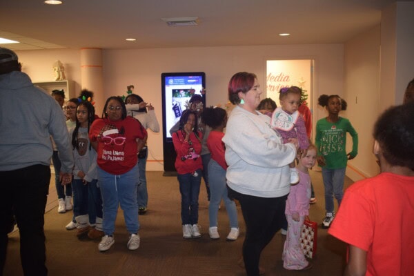 A group of children and adults, some wearing festive holiday clothes, gather in a hallway. One woman holds a small child, while others stand or talk nearby. The atmosphere appears casual and cheerful.