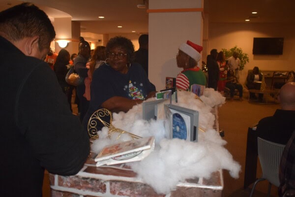 A festive indoor gathering with people socializing. Fake snow and holiday books decorate a brick-patterned counter. One person wears an elf hat and holiday sweater; others are chatting nearby. Warm lighting fills the room.