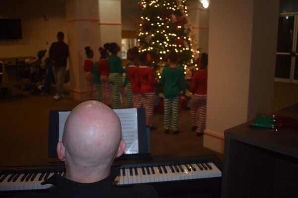 A bald person plays piano while children in striped holiday pajamas stand near a decorated Christmas tree with lights inside a room.