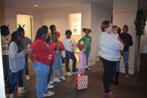 A group of children and a few adults, some in festive attire, stand indoors together. One woman holds a small child, while another child holds a gift bag. Holiday decorations are visible in the background.