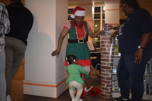 A woman dressed as a festive elf, wearing a Santa hat, smiles and holds hands with a small child in green clothing, while another woman stands nearby, also smiling, in a warmly lit indoor setting.