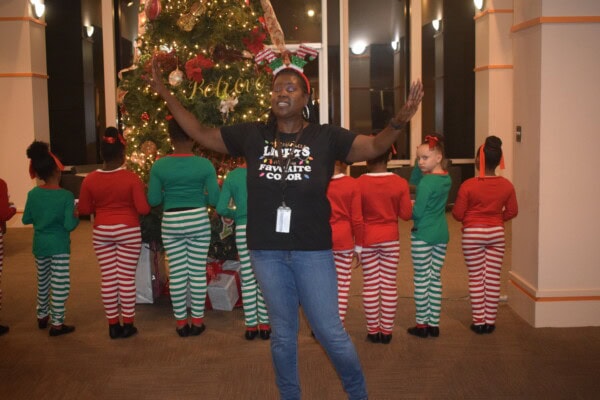 A woman wearing a Black is my favorite color shirt and holiday headband stands with arms raised in front of a Christmas tree, while children in festive red and green outfits face away from the camera.