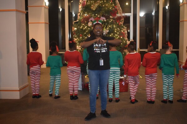 A woman stands with arms crossed in front of her chest facing the camera, while seven children in red, green, and white striped pajamas stand with their backs to the camera in front of a decorated Christmas tree.