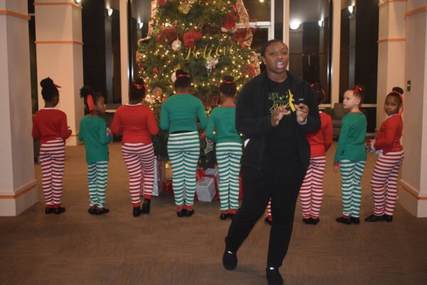 A group of children in red and green striped pajamas stand with their backs to a decorated Christmas tree, while an adult stands in front of them, speaking or singing.