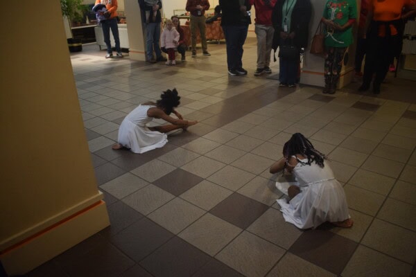Two young girls in white dresses sit on a tiled floor, bending forward with heads down, performing a dance as a group of people watch from the background.